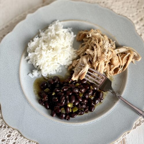 Juicy shredded chicken, white rice, black beans, and a fork on a pastel blue plate, served on a woven tablecloth. Perfect comfort food and easy dinner idea, nutritious and delicious meal for the family.