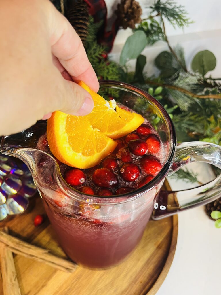 Adding oranges to the Christmas punch in a glass pitcher on a brown tray on the counter