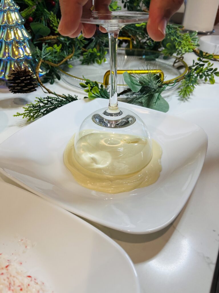 dipping a coupe glass into melted white chocolate on a white plate on at the counter.