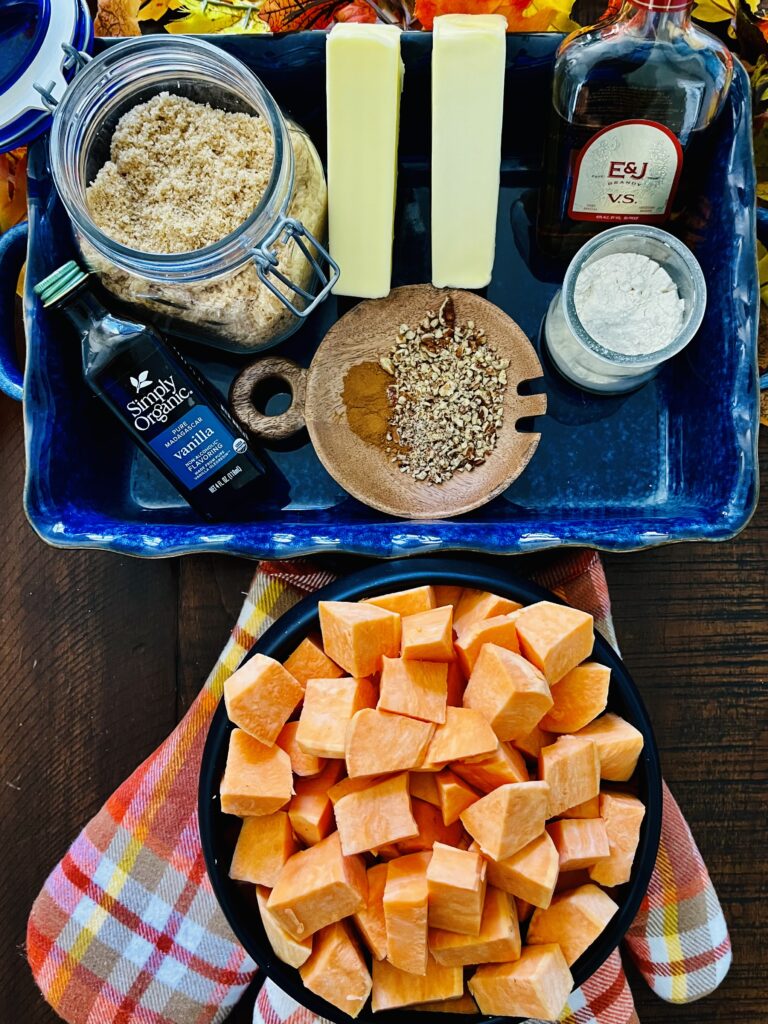 sweet potatoes, vanilla extract, light brown sugar, flour, cinnamon, salted and unsalted butter, brandy and crushed pecans in a blue casserole dish on a wooden table.