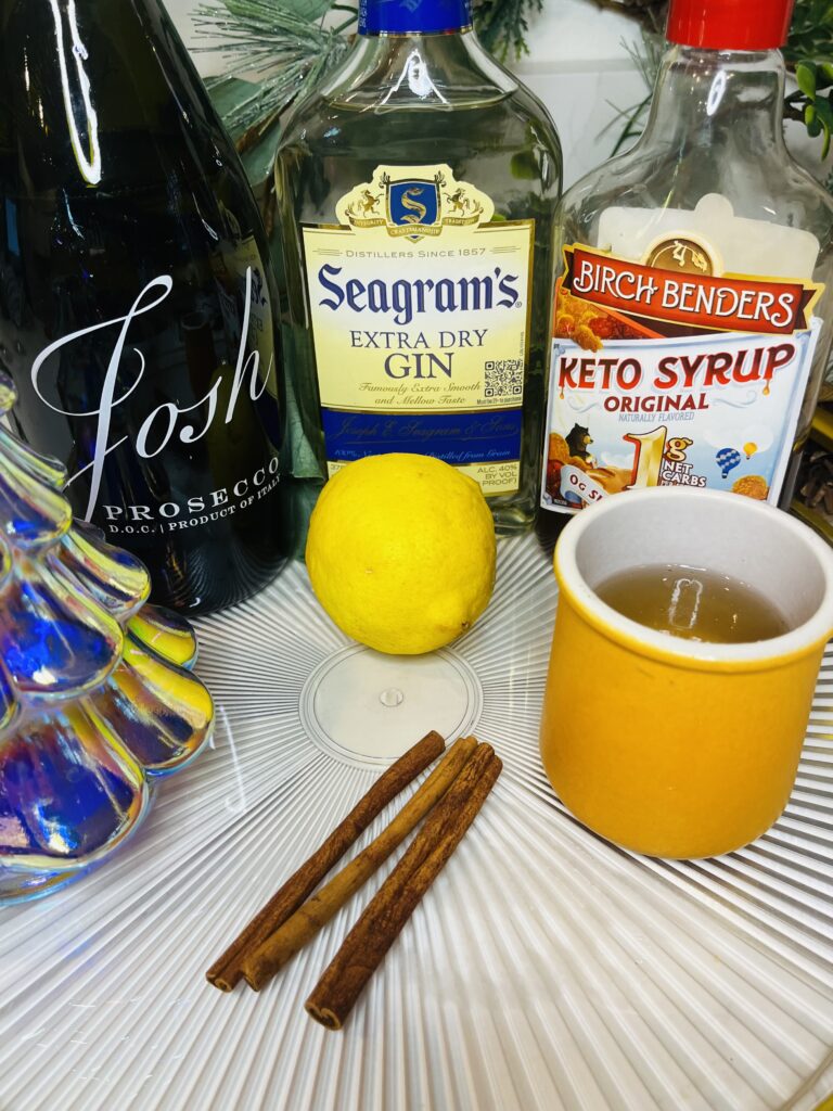 cinnamon simple syrup, keto syrup, gin, lemon , cinnamon sticks and Prosecco on a glass tray on the counter.