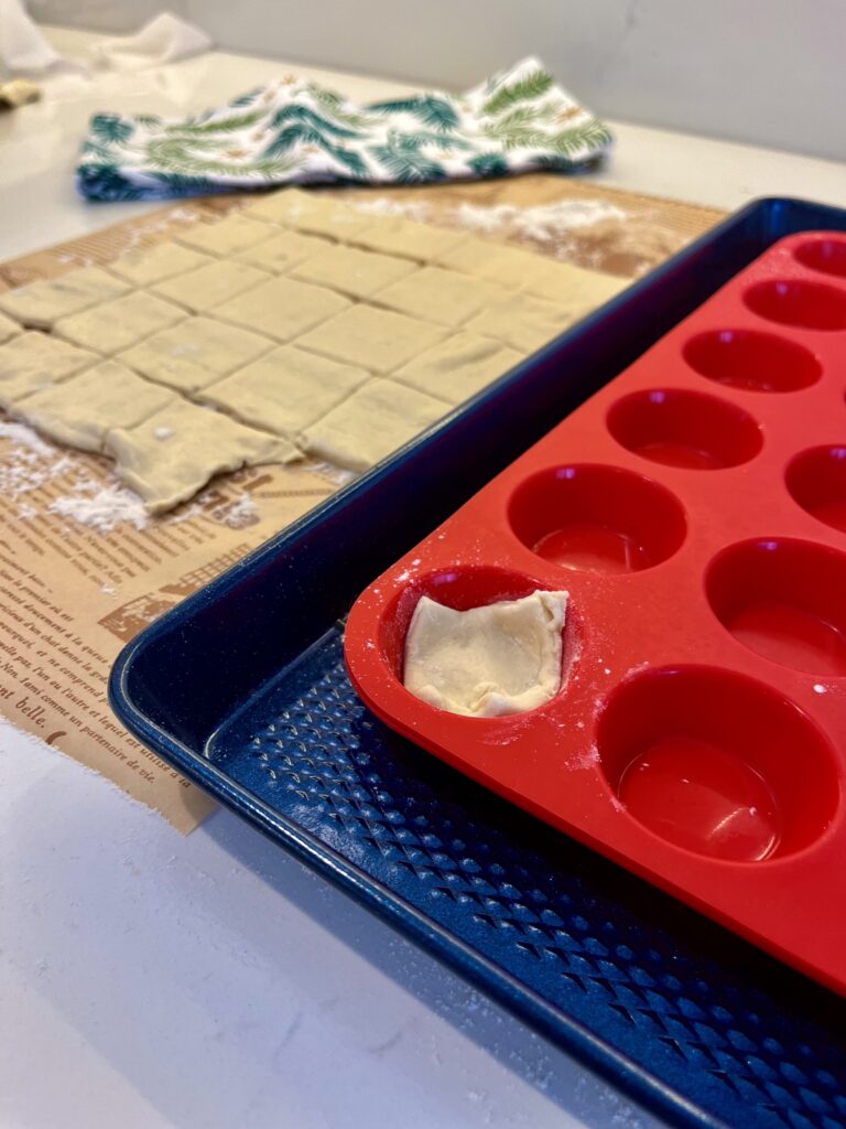 Pastry cut up in 24 pieces on floured parchment paper on the counter and one piece is in the muffin tray on a blue baking sheet on the counter.