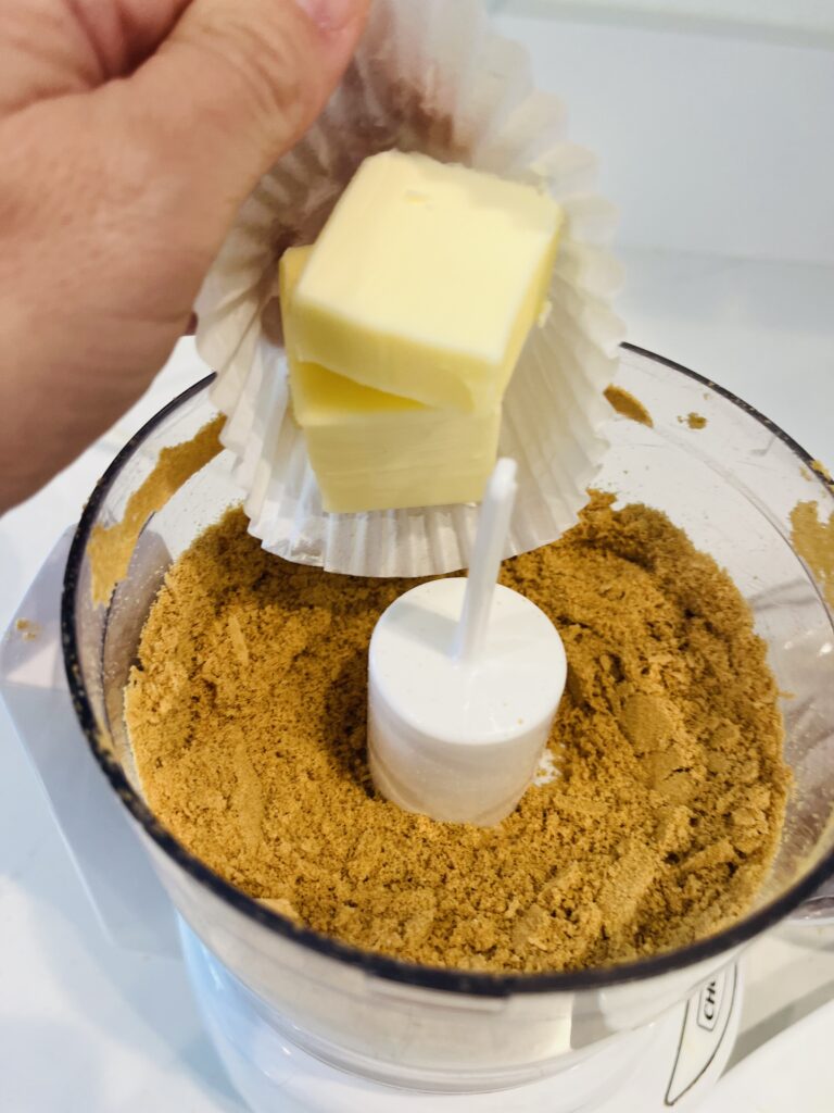 ground up ginger cookies in a food processor. Butter about to be place in the food processor that is on a white kitchen counter.