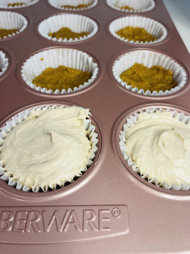 Gingerbread cheesecake filling in muffin tin paper cups in a pink muffin tin on the counter. Some cups have only gingerbread crust-no filling yet.