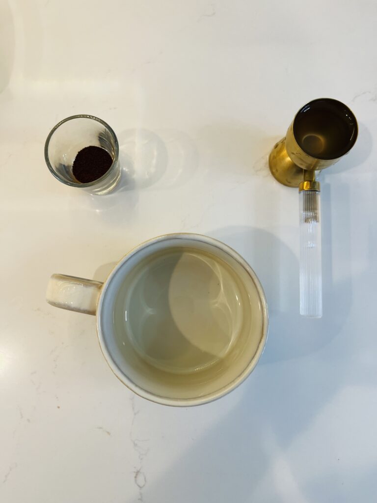 instant espresso a shot glass, an empty cup and water in a gold colored jigger with water in it, on the counter 