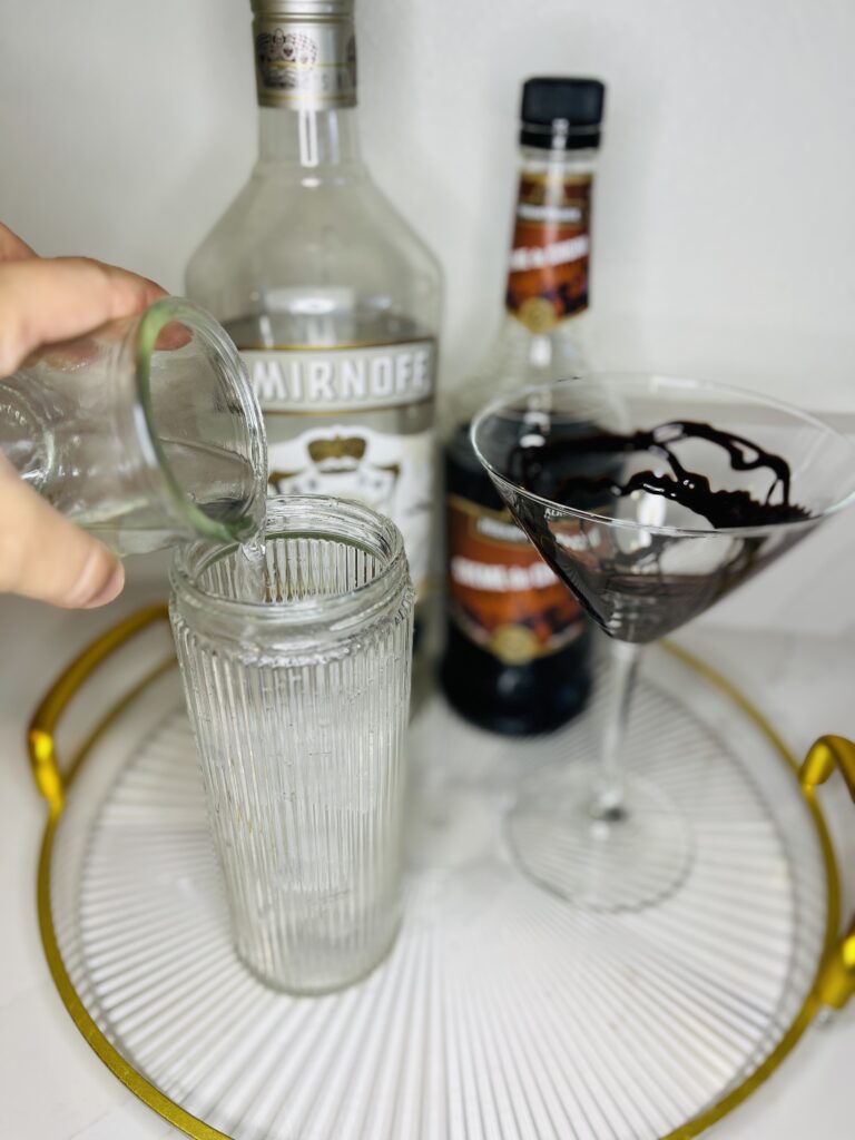 cocktail shaker with vodka being poured into it. Chocolate syrup drizzled martini glass, bottles of vanilla vodka and creme de cacao on a glass tray on the counter.