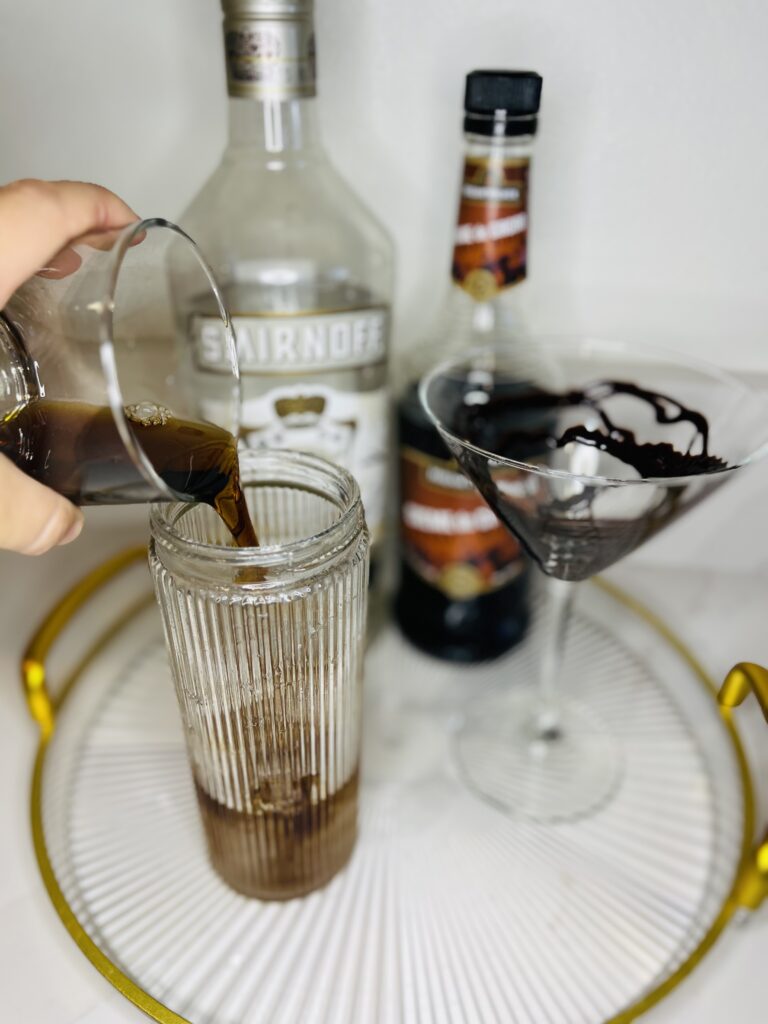 cocktail shaker with creme de cacao ingredient being poured into the cocktail shaker. chocolate syrup drizzled martini glass, bottles of vanilla vodka and creme de cacao on a glass tray on the counter.