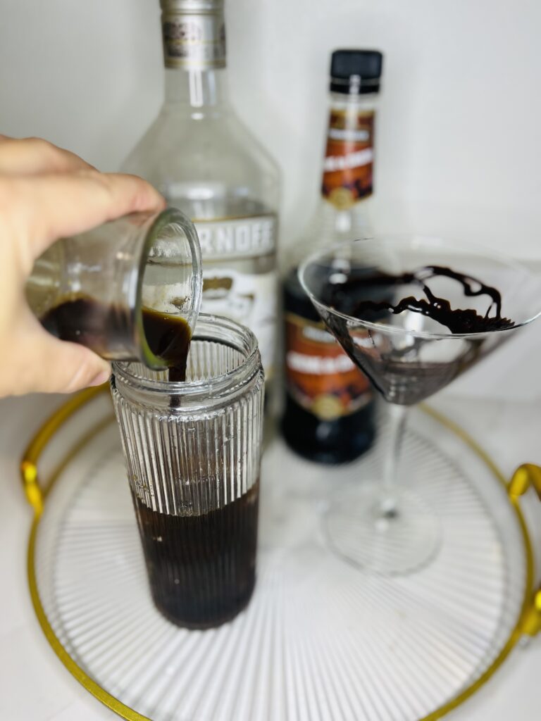 cocktail shaker instant espresso being poured into a cocktail shaker. chocolate syrup drizzled martini glass, vanilla vodka and creme de cacao on a glass tray on the counter.