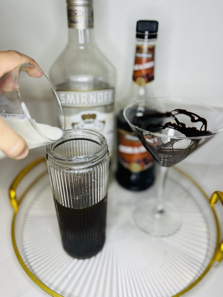 cocktail shaker with chocolate espresso martini ingredients in it. Pouring half and half into the cocktail shaker. A chocolate syrup drizzled martini glass, bottles of vanilla vodka and creme de cacao on a glass tray on the counter.