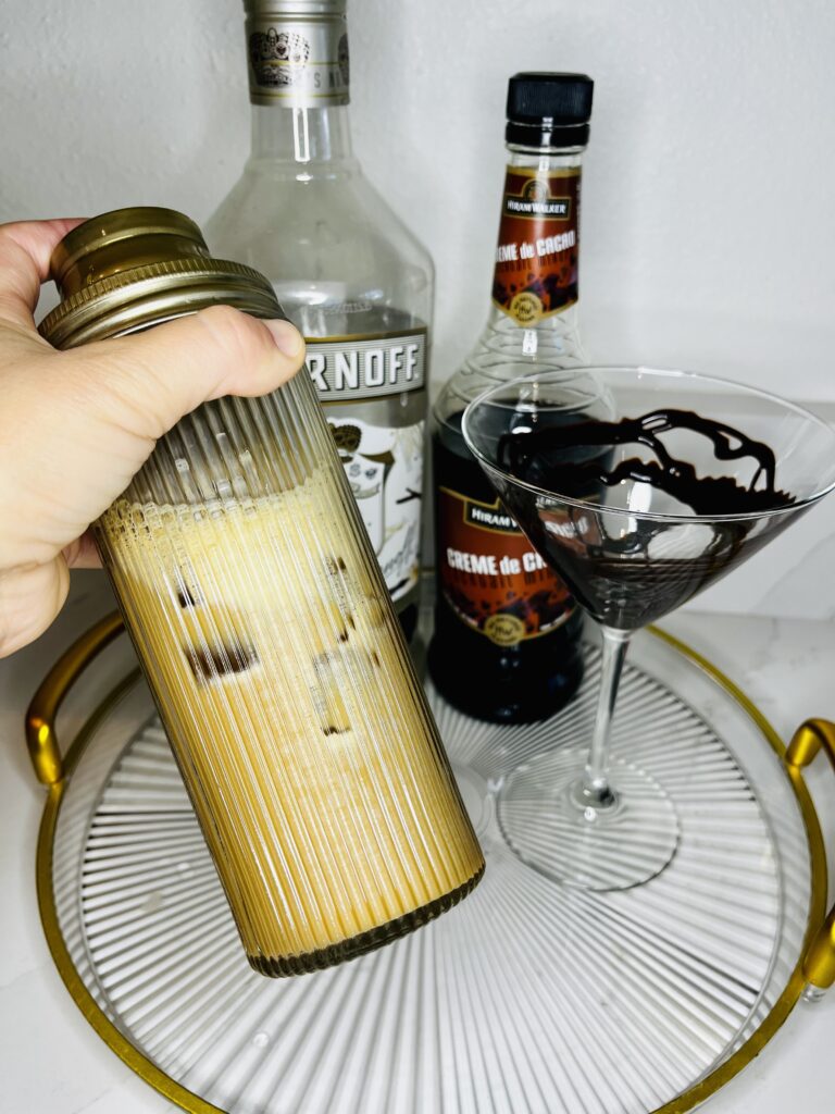 cocktail shaker with chocolate espresso martini ingredients in it. chocolate syrup drizzled martini glass, bottles of vanilla vodka and creme de cacao on a glass tray on the counter.