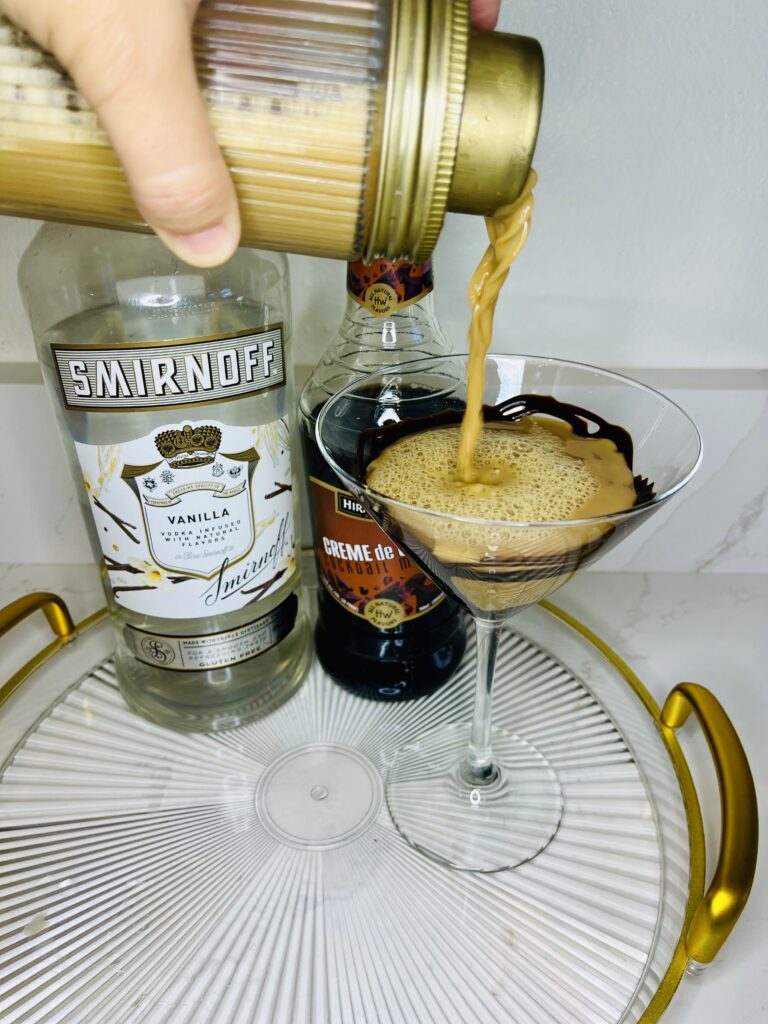 pouring chocolate espresso martini into a martini glass on a glass tray on the counter. Bottles of vanilla vodka and creme de cacao on a glass tray on the counter