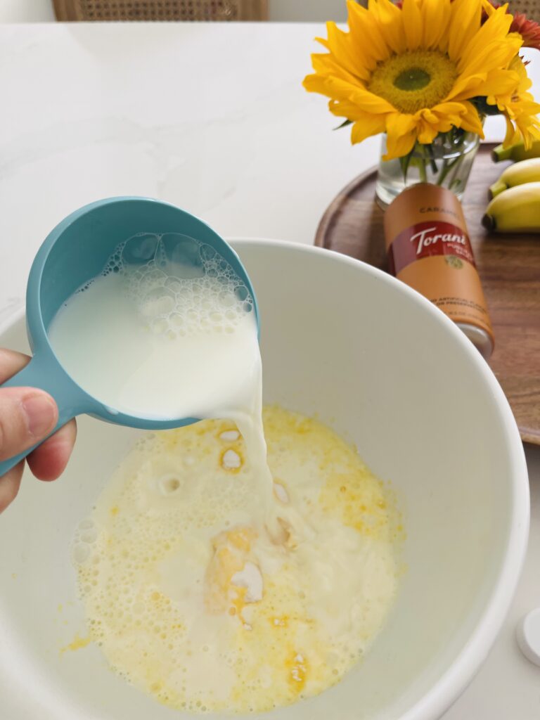 Milk being poured in instant pudding in a large white mixing bowl on the counter. caramel sauce and flowers on a brown tray on the counter.