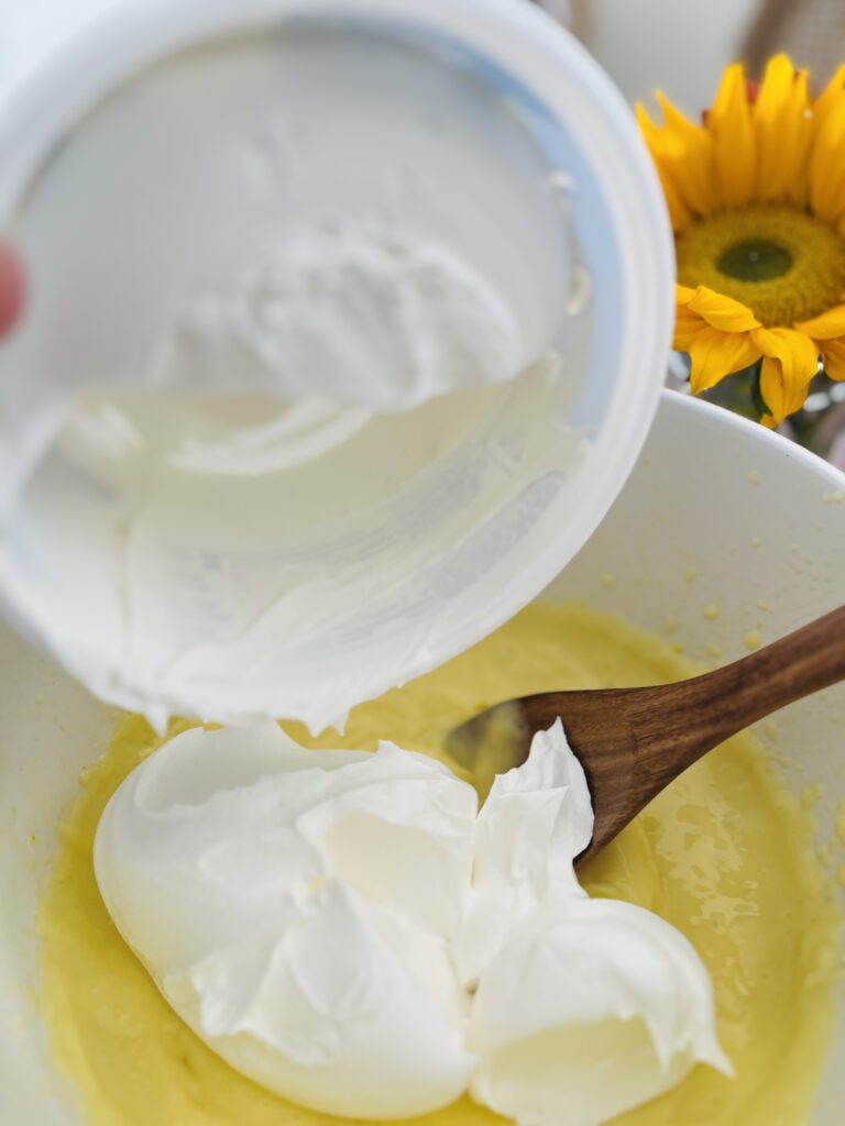 Cool Whip in a container being poured into the vanilla instant pudding in a large mixing bowl on the counter.