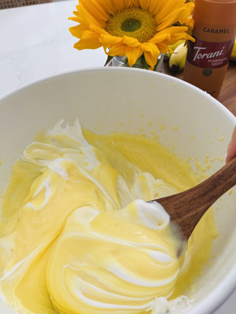 wooden spoon mixing Cool Whip and vanilla pudding in a large mixing bowl on the counter. sunflower and caramel sauce on a brown tray on the counter.