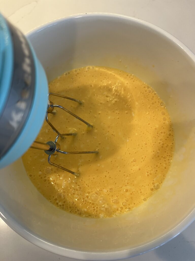 using an electric hand mixer to blend wet ingredients in a large white bowl on the counter