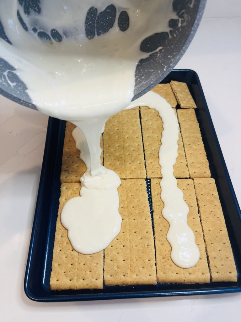 Pouring from a cooking pot the melted marshmallow and butter mixture over the graham crackers on a blue baking sheet on the counter.