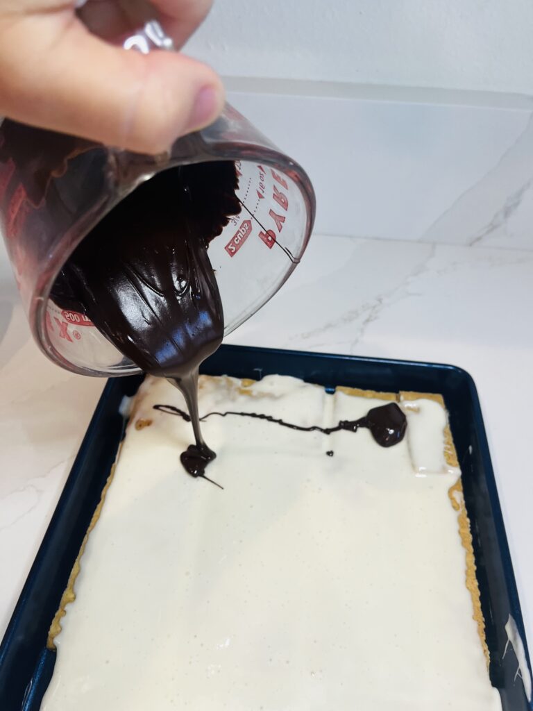 Pouring melted chocolate onto marshmallow layer and graham crackers in a blue baking sheet on the white counter.