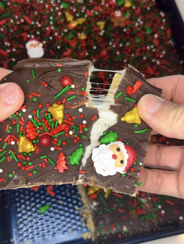 a Christmas s'more being pulled apart. tray of Christmas s'mores in the background on a blue plate on the counter 