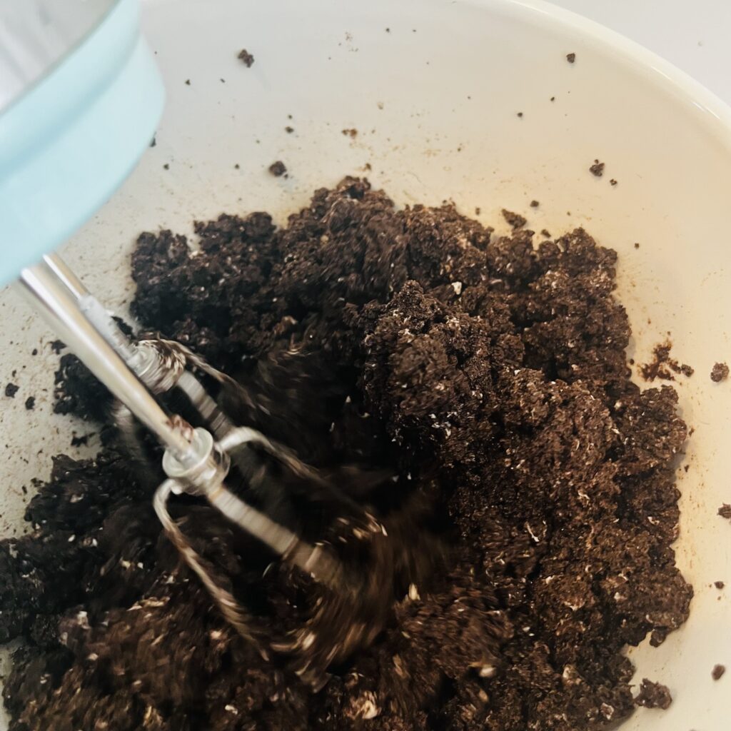 Electric hand mixer mixing Oreo crumbs and cream cheese together in a white mixing bowl on the counter 