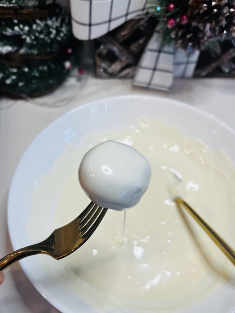 Dipped Oreo Ball into melted white chocolate. A white bowl on the counter. Christmas decorations on the counter.