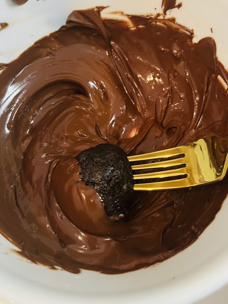 Dipping Oreo Ball into melted semi-sweet chocolate with a fork into a white bowl, on the counter.