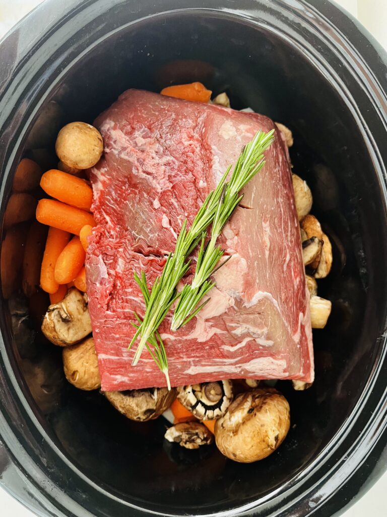 Raw beef pot roast, mushrooms, carrots, chopped onion and rosemary sprigs in a black slow cooker on the counter. 