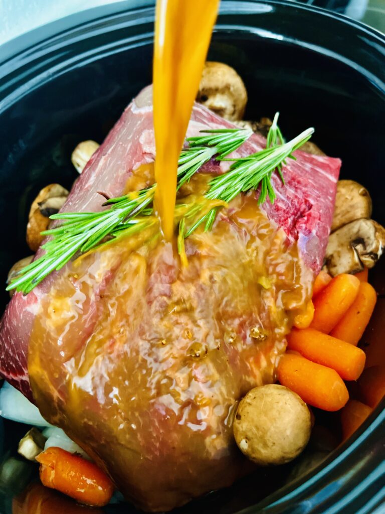 Raw beef pot roast with seasoning and water being poured over it,mushrooms, carrots, chopped onion and rosemary sprigs in a black slow cooker on the counter. 