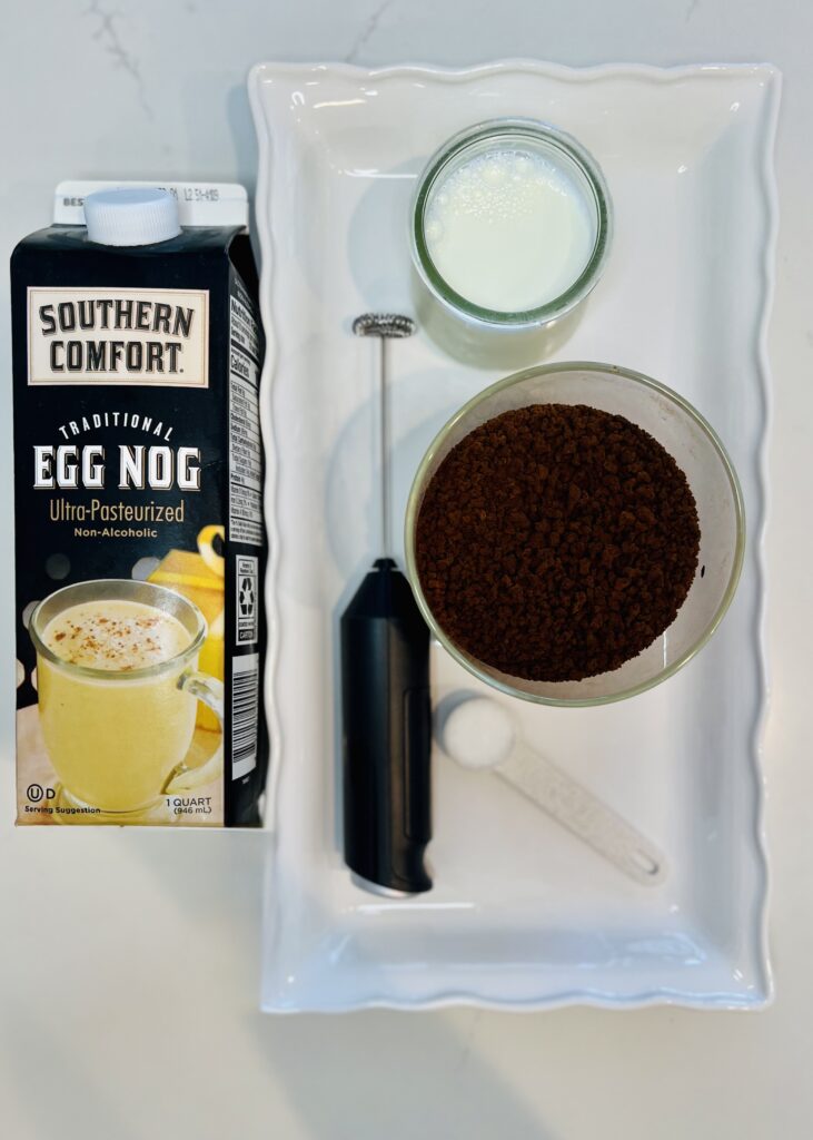 Eggnog in a carton on the counter. Milk in a glass container, instant coffee in a glass container, teaspoon of sugar, and a milk frother on a white tray on the counter.