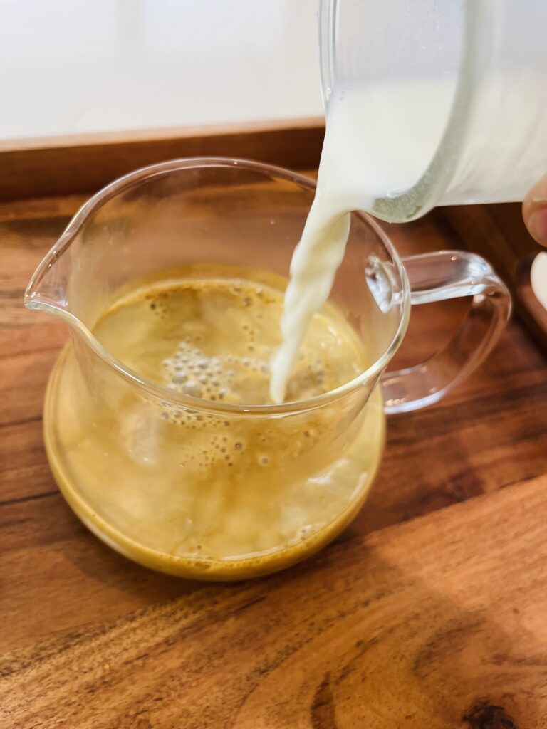 pouring whole milk into a glass with a spout with the sugar and instant coffee on a brown tray on the counter.