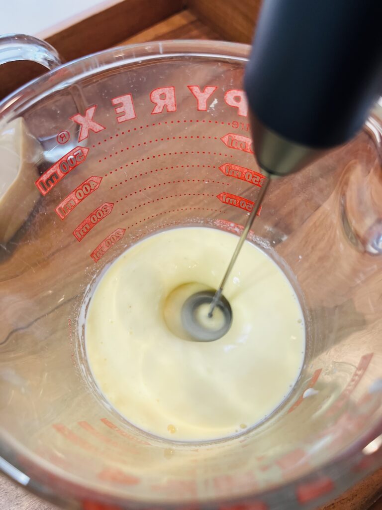 using a frother to whip and froth eggnog in a glass measuring cup on a brown tray.