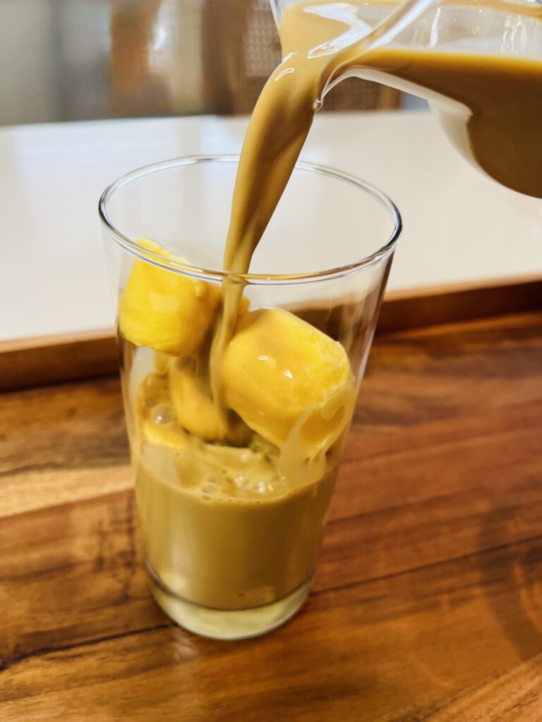 Pouring coffee and milk over eggnog ice cubes in a tall glass on a brown tray on the counter.