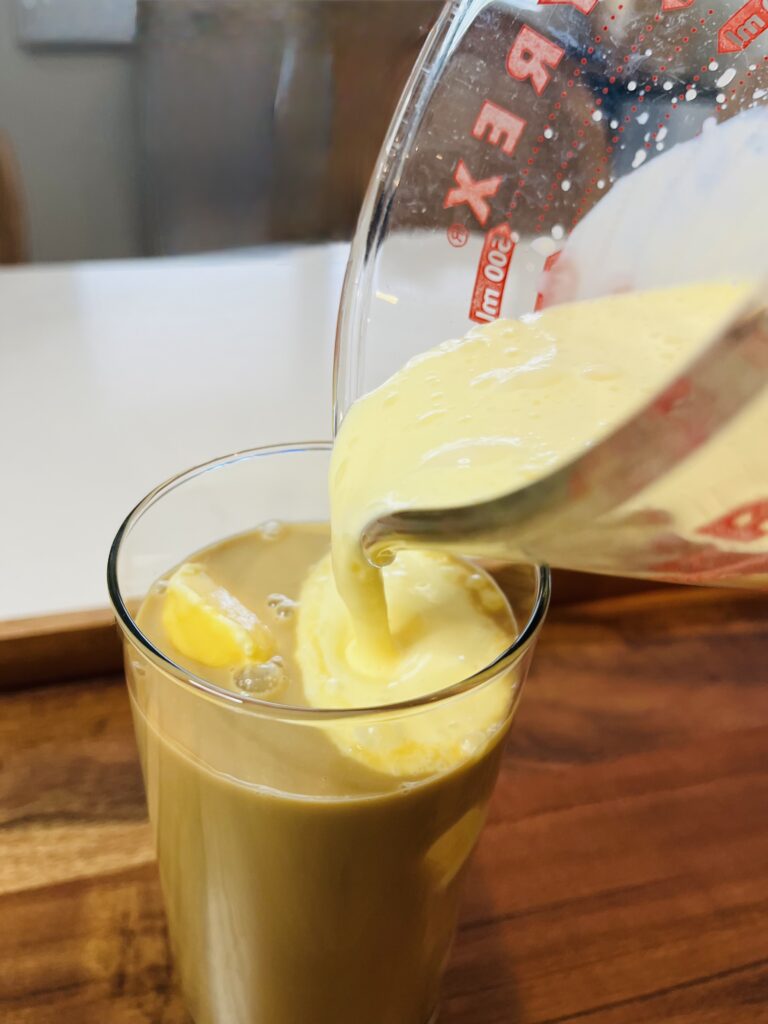 pouring whipped eggnog over coffee mixture in a tall glass on a brown tray on the counter.