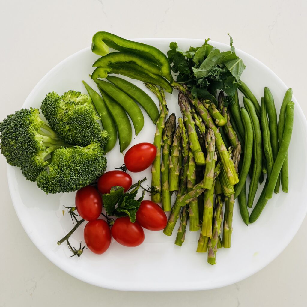 broccoli, snow peas, asparagus, sugar snap peas or snow peas, cherry or grape tomatoes, green beans, green pepper, kale and fresh mint leaves on a white plate on the counter.