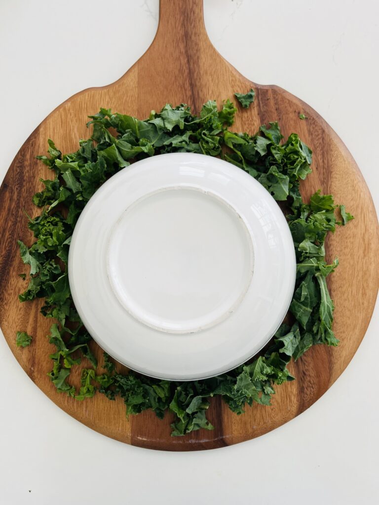 Kale in a wreath shape on a brown board with a white upside down bowl in the middle on the counter.