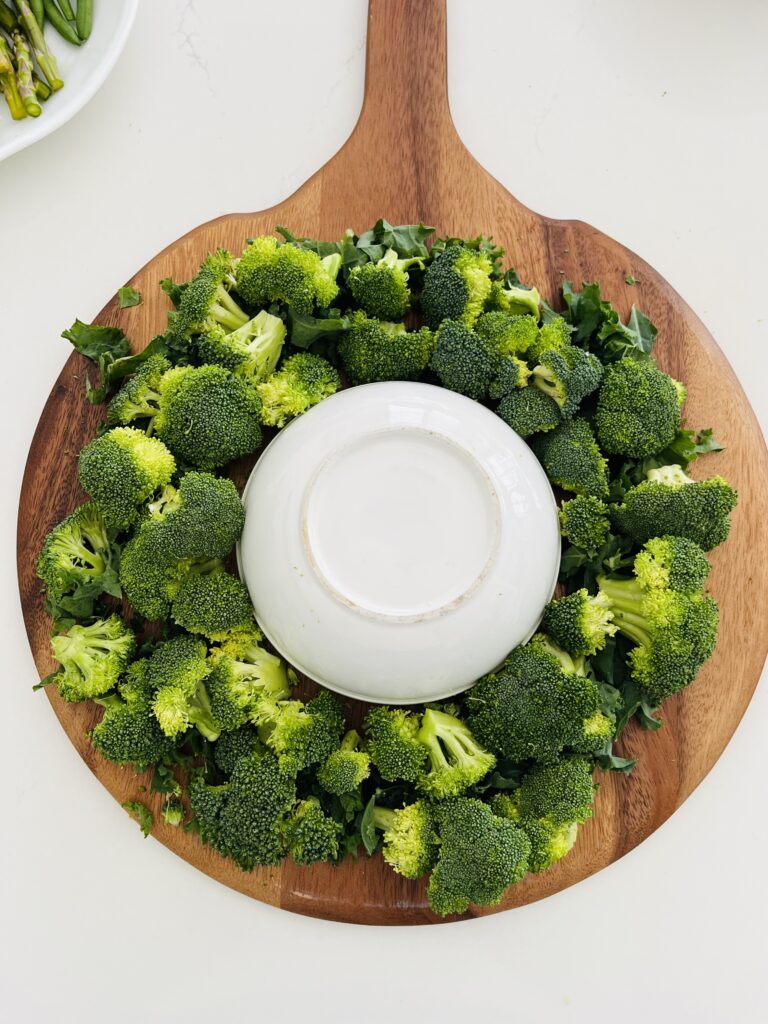 Kale and broccoli  in a wreath shape on a brown board with a white upside down bowl in the middle on the counter.