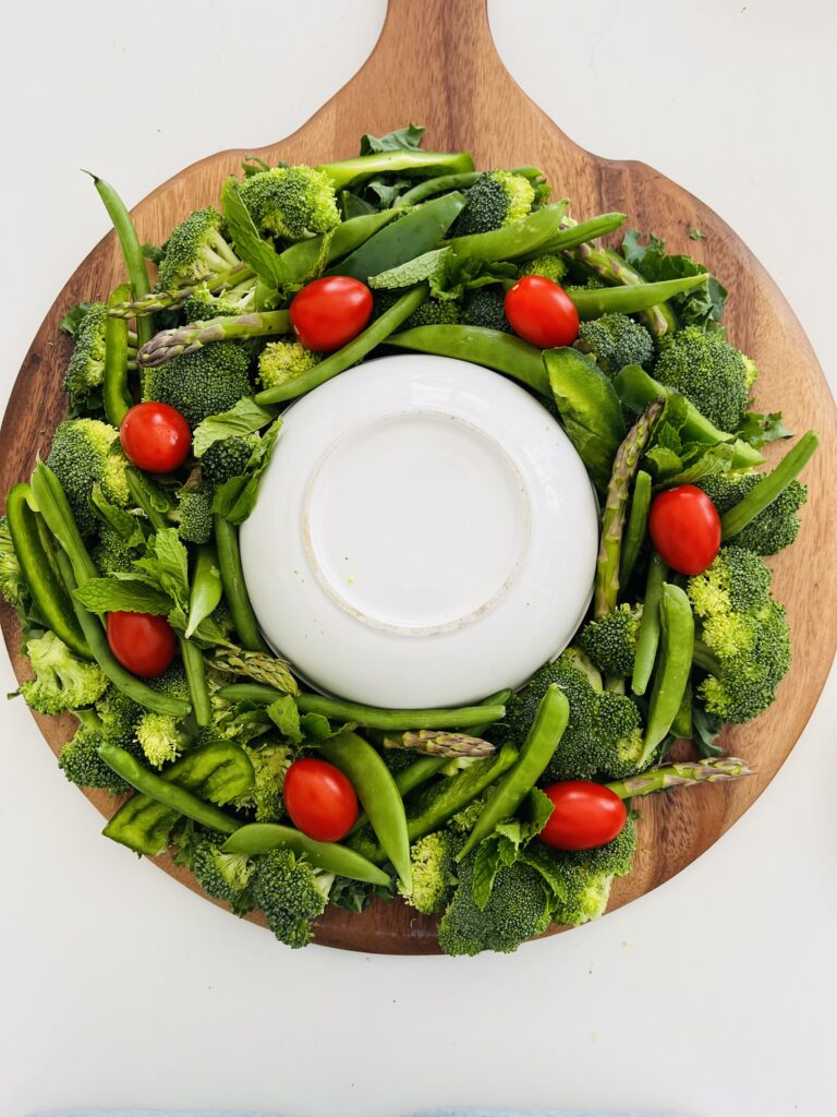 Green vegetables in a wreath shape on a brown board with a white upside down bowl in the middle on the counter.