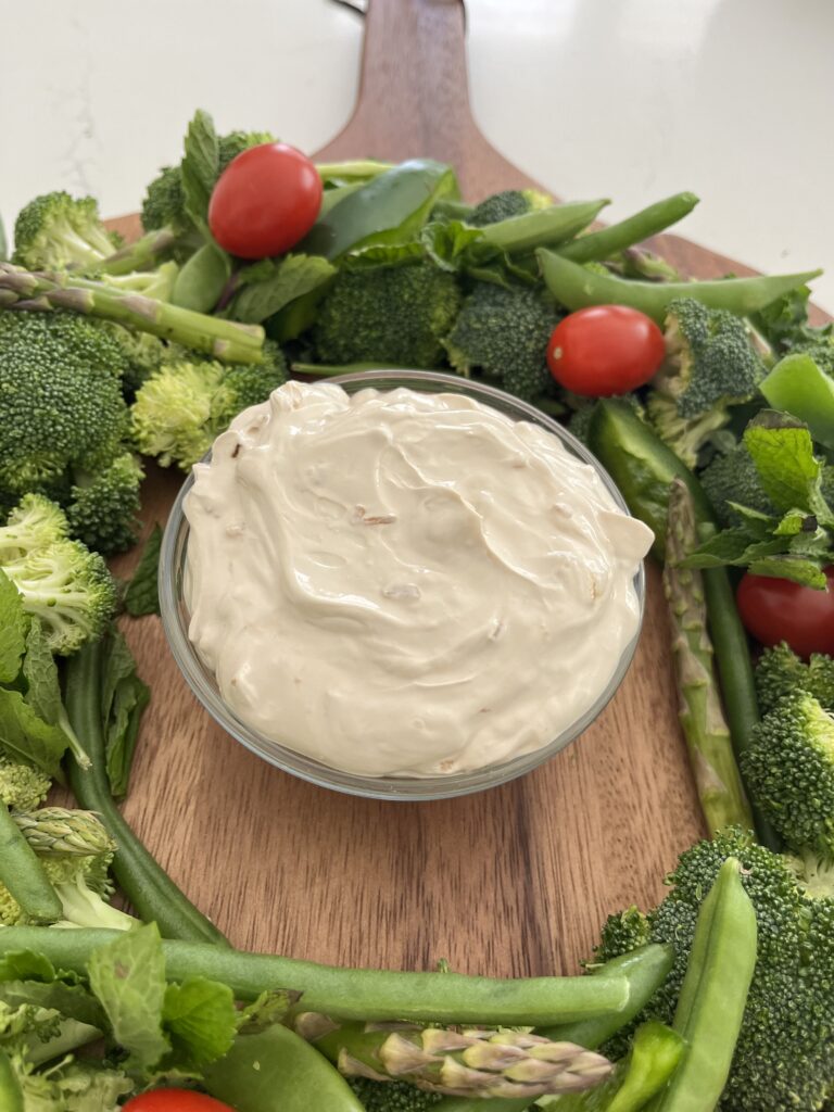 Onion dip in a glass bowl surrounded by assorted vegetables on a brown platter on the counter.