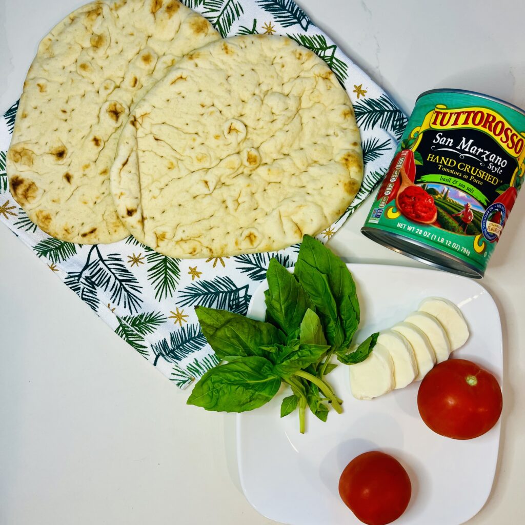 Naan bread, can of hand crushed san Marzano tomatoes, 2 roma tomatoes, sliced mozzarella cheese and fresh basil on a white plate on the counter.