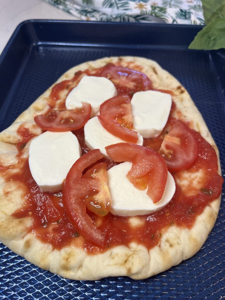 naan bread with tomato sauce, mozzarella cheese and a thinly sliced tomato on a blue baking sheet on the counter.