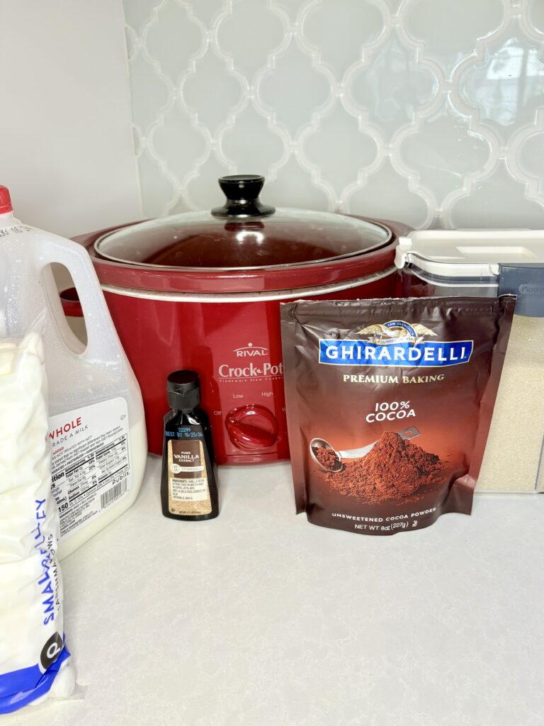 a slow cooker on a counter with cocoa powder, milk, sugar, vanilla extract and marshmallows