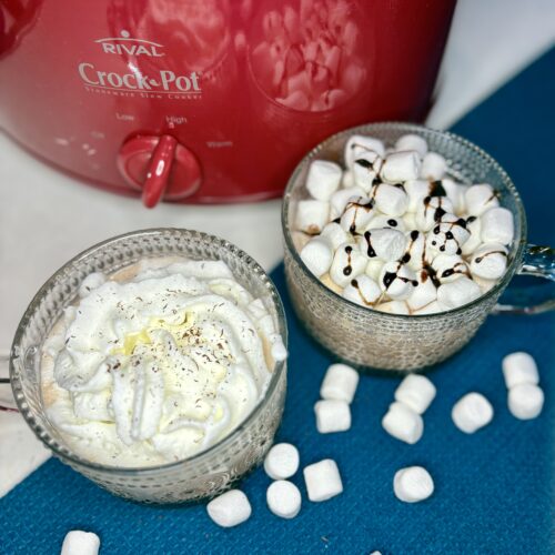 2 mugs of hot cocoa on a counter with a slow cooker