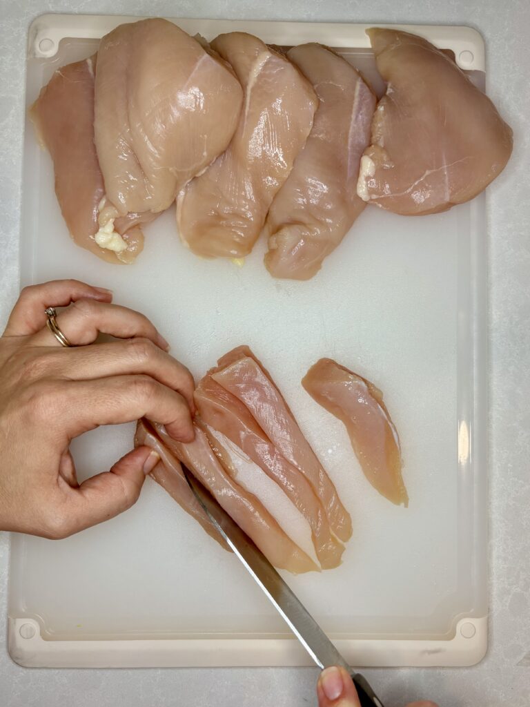 raw chicken breasts on a cutting board being sliced into strips