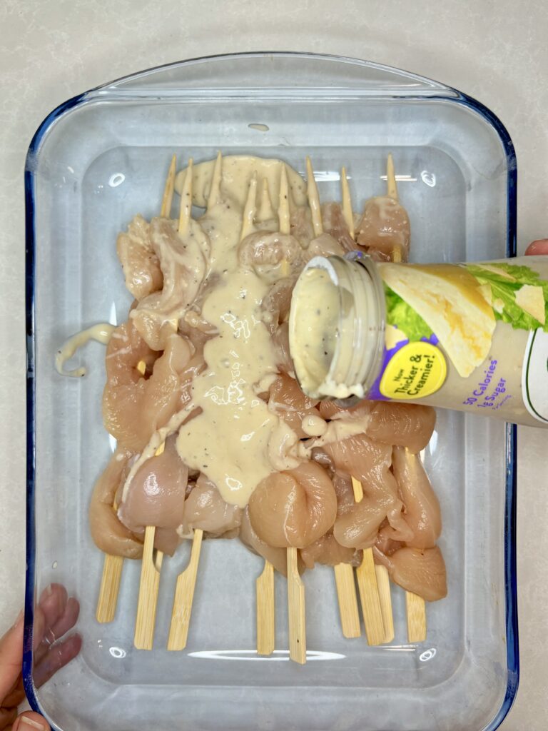 caesar dressing being poured over raw chicken skewers in a dish