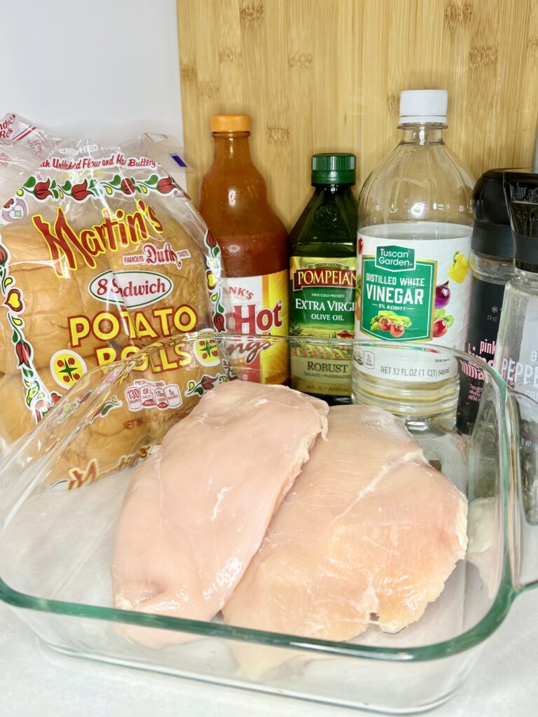 raw chicken breasts, vinegar, oil, buffalo sauce, salt, pepper and buns on a counter