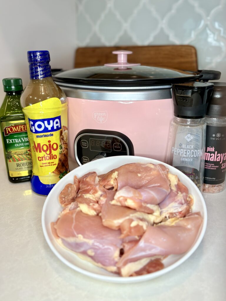 raw chicken thighs on a plate on a counter next to a slow cooker, mojo criollo marinade, olive oil, salt, and pepper