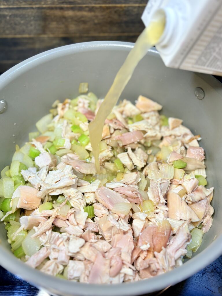 broth being poured in to a pot with chicken, celery, onion, and garlic