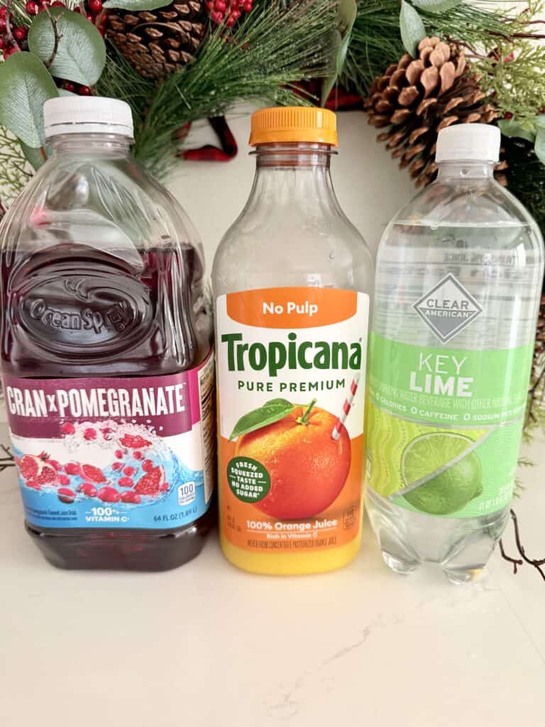 Three bottles of juice, including cranberry pomegranate, orange, and key lime, are arranged on a table with holiday decorations in the background.