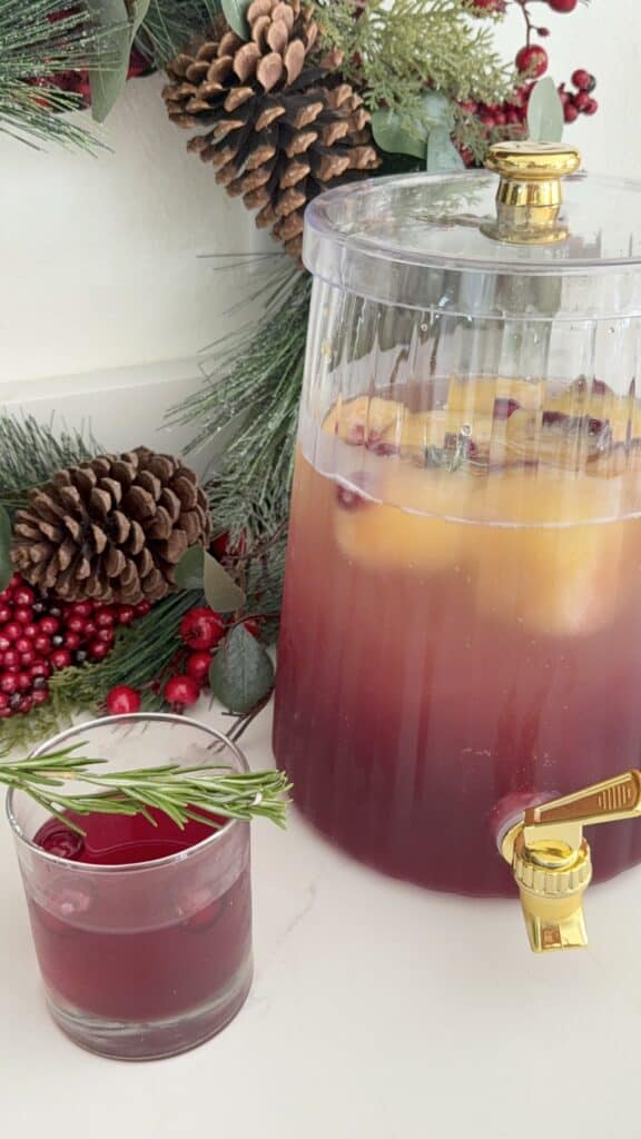 Refreshing sparkling cranberry mocktail in a glass dispenser with a spigot, garnished with fresh cranberries and holiday greenery.