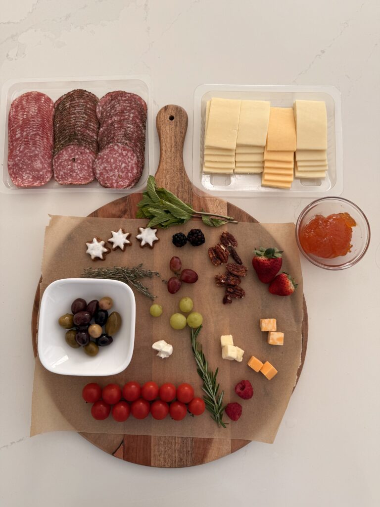 assorted thinly sliced salami, assorted sliced cheeses, assorted cubed cheeses, mozzarella pearls, apricot jam, olives, cherry tomatoes, assorted berries, white and red grapes, assorted nuts, cookies of choice, crackers of choices, sprigs of rosemary, thyme and mint herbs for decorating on a large brown board on the counter.