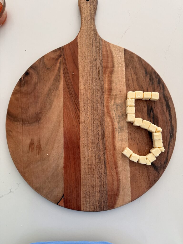 Cubed cheeses making the shape of the number five on the large brown board on the counter.